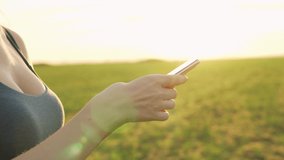Athletic Girl Uses Smartphone In Summer Field Park After Jogging At Sunset. Close-up of Hands Fingers and Cell Phone, Side View. SMS, Chatting, Communication, Social Networks, Video Viewing, Online.