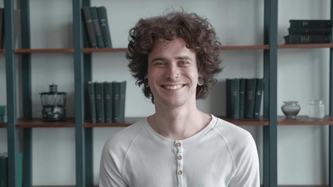 Young millennial with curly hair and pierced in the nose handsome with ears tunnels, and smiling joyfully of library interior background. Close up of stylish man.
