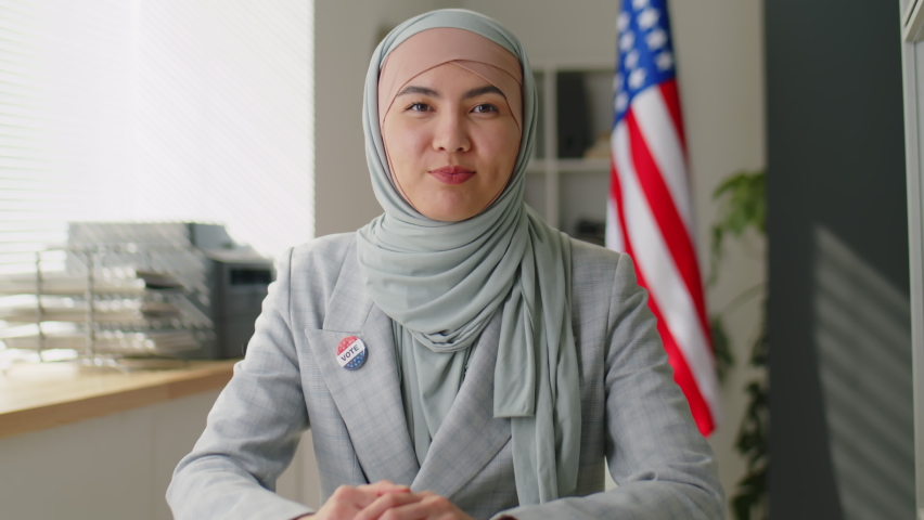 Young muslim woman in hijab with Vote badge speaking on camera in office with U.S. national flag while inviting people to participate in election