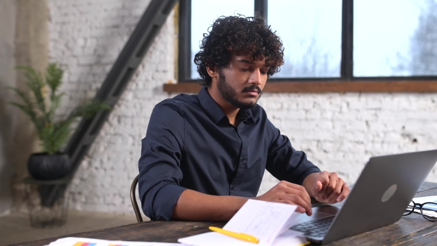 Front view of positive Hindi man in smart casual shirt using laptop while sitting at the desk in his flat. Young Indian male student watching webinars, educational courses, learning on the distance Royalty-Free Stock Footage #1091516237