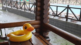A yellow inflatable duck, lies on massive wooden table in cozy gazebo of one of houses at recreation center against the backdrop of large pool covered with heavy pouring rain. 4K UHD slow-mo video
