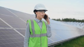 Beautiful caucasian female worker in signal vest and protective helmet standing among solar panels and talking on mobile phone.
