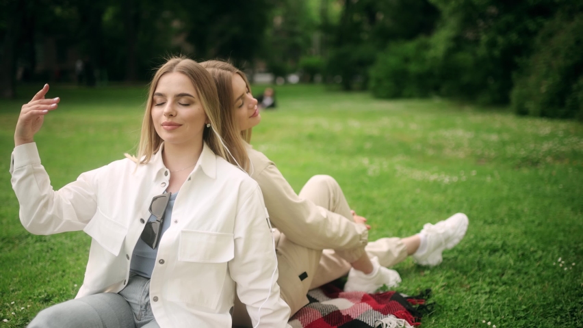 Two beautiful blonde young women listen to music in the park. Sitting back to back