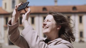 Delighted, happy young woman with curly hair takes pictures, shoots video on phone in horizontal orientation in courtyard of restored medieval castle. Sunny spring day, wind blows on hair.