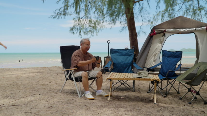 elderly enjoy with camera on the beach vacation time. Asian senior sitting on the beach with family holding camera looking picture near tents, old man holiday camping with family beach background.