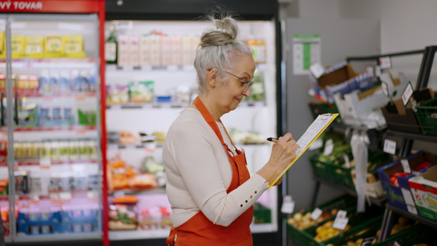 Confident senior shop assistant in supermarket in vegetable shell, in bakcground is her colleague filling stock.