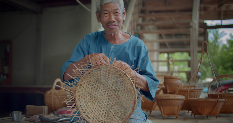 Slow motion scene of a happy smiling Asian male farmer who is an elderly native basketry artisan weave basket from bamboo that are cut into strips for weaving folk crafts in rural areas of Thailand. Royalty-Free Stock Footage #1091531243