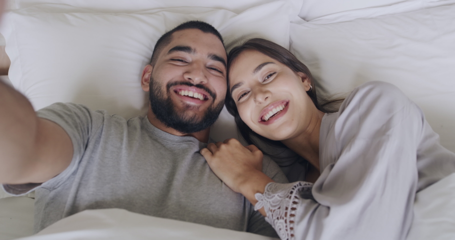 Goofy young interracial couple making funny faces and being silly while taking a selfie in bed. Playful lovers laughing and having fun while laying together. Sticking out tongue and looking cross eyed | Shutterstock HD Video #1091532675