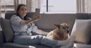 Happy young woman using her smartphone for a video call on the couch and waving at her house. Caucasian woman with her puppy. Fun and relaxing day on the couch with your pet and phone. Love and kiss