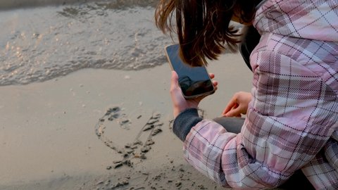 A teenage girl draws a heart on the sand and shoots it on a smartphone. A wave washes away a heart drawn in the sand