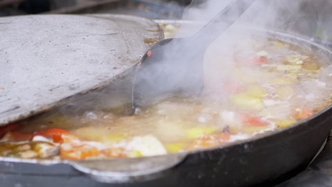 Street Chef Prepares a Vegetable Stew Outdoors in a Cast Iron Skillet or Wok. The cook mixes stewed vegetables with a spoon in boiling bouillain. Food, boiling on steam and smoke. Zoom. Slow motion.