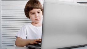 A focused and attentive child sits and does a task on a laptop. Close-up. 5 year old boy solves a test by a distance online education program. Smart kid. White room, furniture, t-shirt. Copy space.