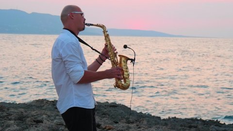 Young slender bald man saxophonist in white shirt plays golden alt saxophone on musical instrument, on seashore, against background of sea, beautiful sunset on of Cyprus. Record sound to microphone.