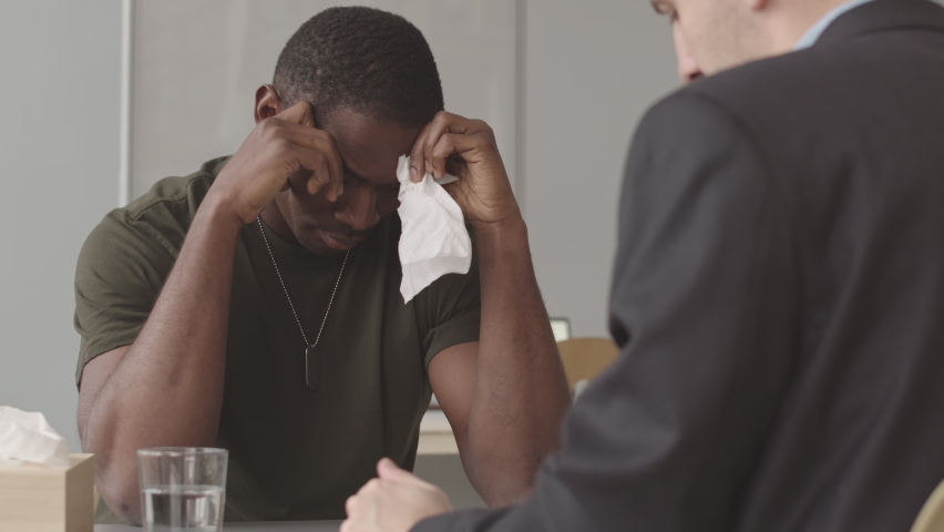 Waist up slowmo of distressed African American soldier with PTSD thinking of horrors of war while having therapy session with professional military psychologist | Shutterstock HD Video #1091557007
