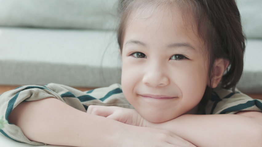 Face close up of adorable asian girl in comfortable living room at home looks at the camera with smiling and shows positive emotion, enjoyment, happiness of child with beautiful sunlight.