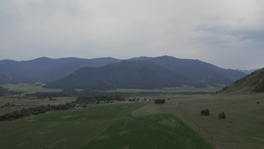 Rural fields and mountains under dramatic sky with white clouds in Altai, Siberia, Russia. Beautiful summer nature landscape at during daytime. Aerial view from a drone