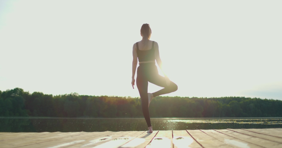 Beautiful young woman practices yoga asana tree pose on the wooden deck near the lake. Rear view. Slow motion | Shutterstock HD Video #1091563161