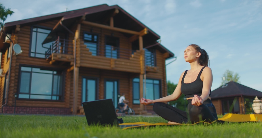 Young woman meditates with laptop in the backyard on the green grass against the background of country house. Slow motion | Shutterstock HD Video #1091563743