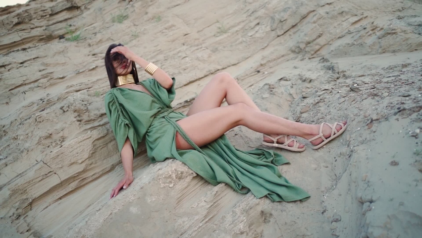 Perfect Brunette Model with Long Hair in Oriental Style Clothes and a Gold Jewelry around Her Neck Poses against the Backdrop Sand on Bright Sunny Day. Beautiful Woman at the Photo Shoot. Slow motion