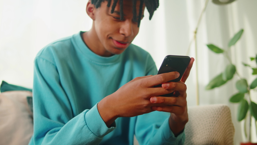 African American man using smartphone. Young guy texting at phone, teenager relaxing at home. Communicating with family and friends online, sitting on sofa in living room. Royalty-Free Stock Footage #1091566135