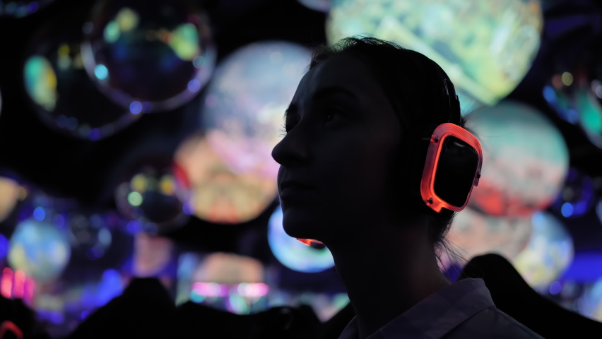 Woman wearing wireless black headphones and looking around in dark room of interactive exhibition or museum with colorful illumination - close up. Futuristic, immersive, entertainment concept Royalty-Free Stock Footage #1091568537
