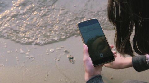 A teenage girl draws a heart on the sand and shoots it on a smartphone. A wave washes away a heart drawn in the sand