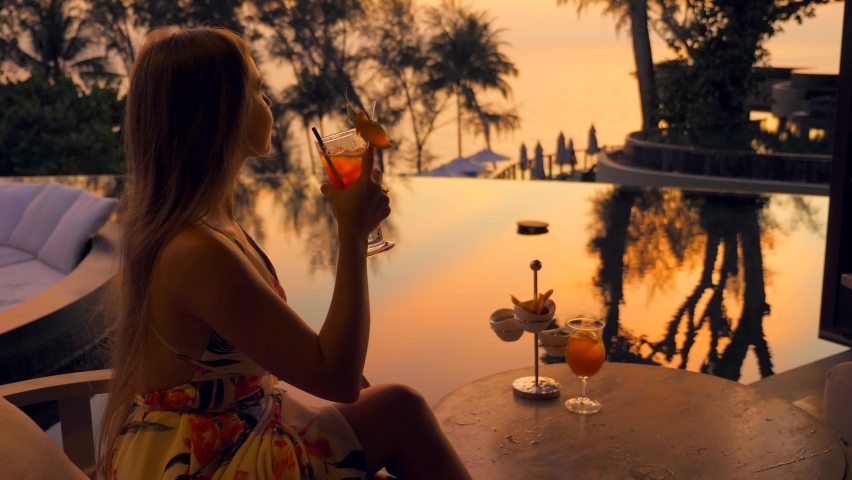 Young attractive woman with cocktail sitting in outdoors restaurant at luxury hotel or resort during sunset time. Summer travel, holidays or vacations concept. Girl in evening gown holding mocktail. | Shutterstock HD Video #1091572289
