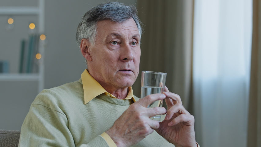 Old sick senior man gray-haired aged grandfather patient pensioner sits in room on couch holds transparent glass with cold clean still water drinks talking in hospital doctor appointment, healthcare Royalty-Free Stock Footage #1091573131