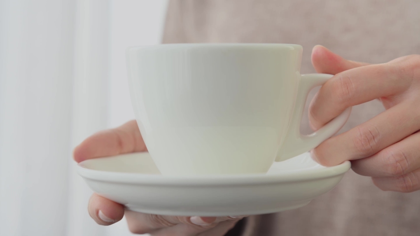 Hands of young woman girl in cozy beige sweater hold white ceramic cup of hot tea or coffee with a saucer near the window. Hands with gentle manicure hold cup close-up | Shutterstock HD Video #1091574743