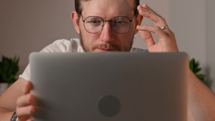 Young bearded caucasian man in computer glasses works on laptop at home. Student, freelancer, businessman surfs the internet watches videos and smiles pretty. Focused guy enjoys work real life concept | Shutterstock HD Video #1091575737