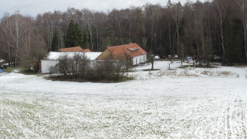 Farmhouse with snowy fields and leafless trees in winter countryside.