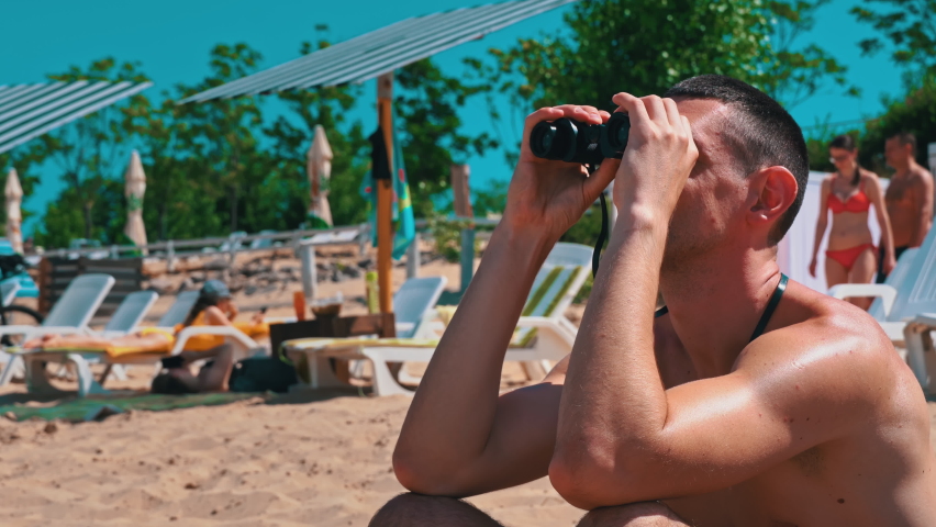 Man on the beach in swimming trunks is watching someone through binoculars. The lifeguard on the beach keeps order in the water. Curious man looks through binoculars at nature. maritime emergency | Shutterstock HD Video #1091583019