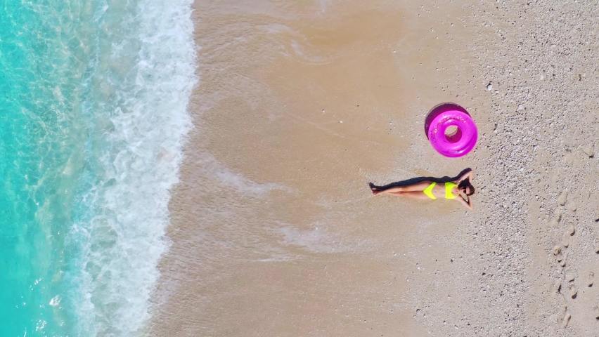 Aerial view of the lying beautiful young woman with pink swim ring on the sandy beach near sea with waves at sunset. Summer vacation in Lefkada island, Greece. Top view from drone. Clear azure water Royalty-Free Stock Footage #1091585249