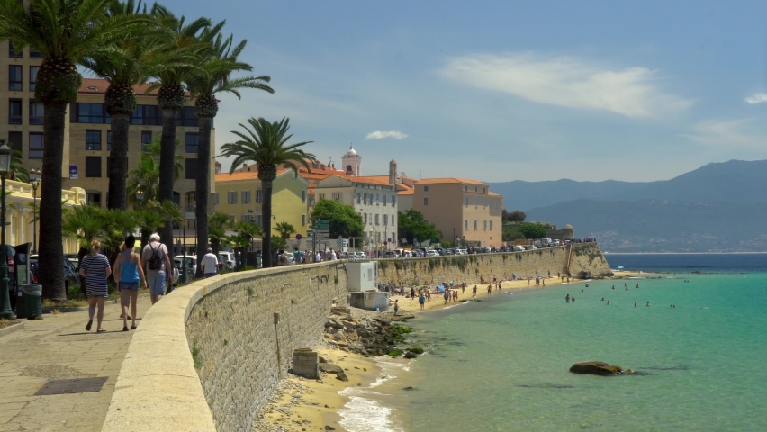 Ajaccio, Corsica, France - June 2022: Water front street with tourists walking, palm trees, wall, buildings, sandy beach. Sunny day, blue sky, turquoise water 