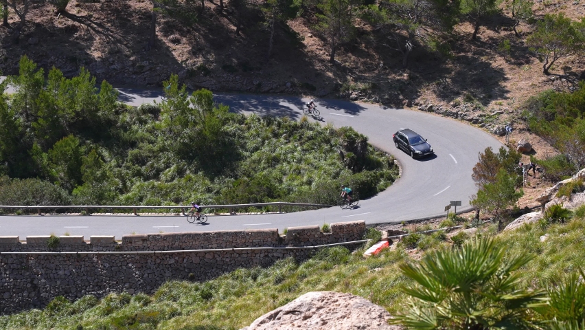 Athletes cycling on winding mountain road. Cyclists riding bicycles and car moving amidst trees. High angle view of bikers training during sunny day on the island of Mallorca.