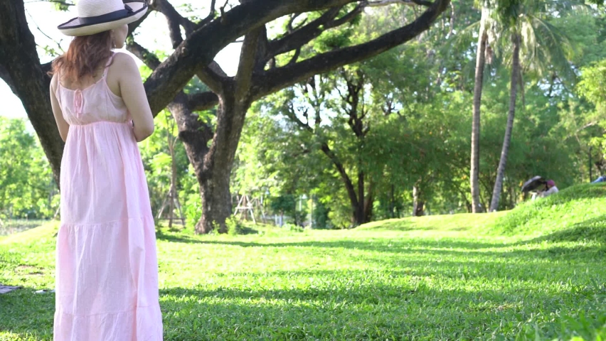 Adult adorable woman wearing long pink loose dress with hat, walks on green fresh grass under the tree at daytime with relax and transquilment