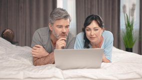 Happy middle aged man and woman using laptop computer at home watching online show lying in bed together. Adult couple looking at gadget screen making video call chatting in bedroom at home. - Powered by Shutterstock - Get 15% off with code: PIKWIZARD15