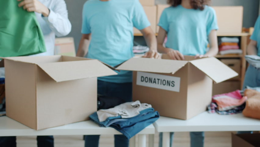 Close-up of boxes for donation and volunteers in uniform packing clothing in office doing charity helping poor and needy. Volunteering and humanitarian aid concept. Royalty-Free Stock Footage #1091600167