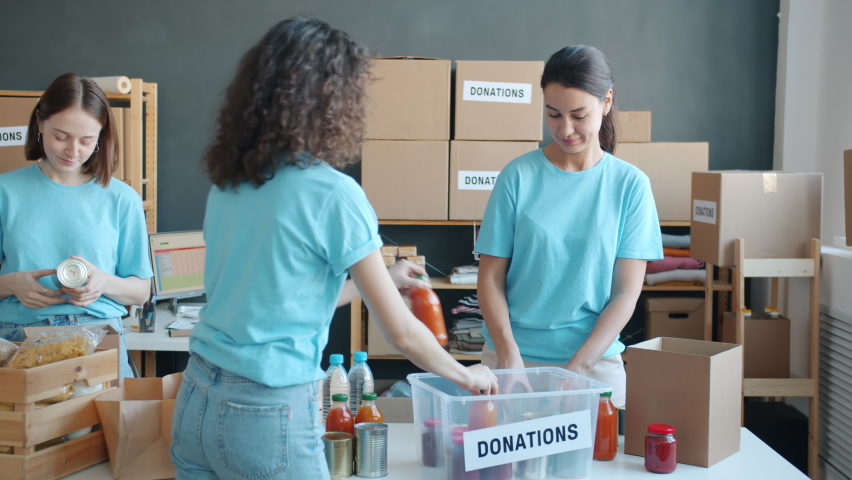 Group of charity company employees packing donation boxes with food to feed poor and needy. Young people in uniform working and talking together. | Shutterstock HD Video #1091600253