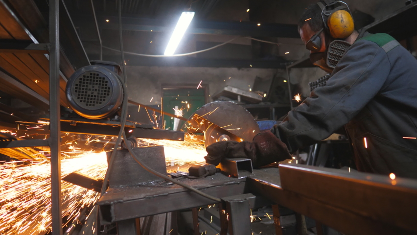 Industrial professional worker use saw cutting machine to cut metal tube. Guy in safety protection wear with circular saw grinding steel in garage. Man sawing metal. Sparks fly from metal. Dolly shot