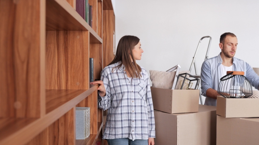 Young couple unpacking cardboard box and putting stuff on the shelving unit in new apartment on moving day. Happy homeowners on relocation day. Real estate and tenancy concept. Young family. Royalty-Free Stock Footage #1091604421