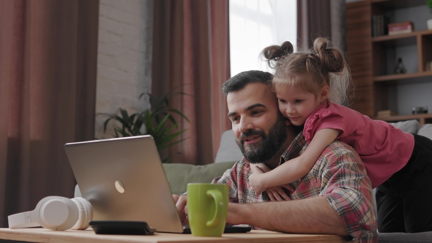 Young handsome man works from home at laptop, focus attention on laptop screen while his little daughter hugging her beloved dad around neck. Family love and family bond concept. Royalty-Free Stock Footage #1091604433