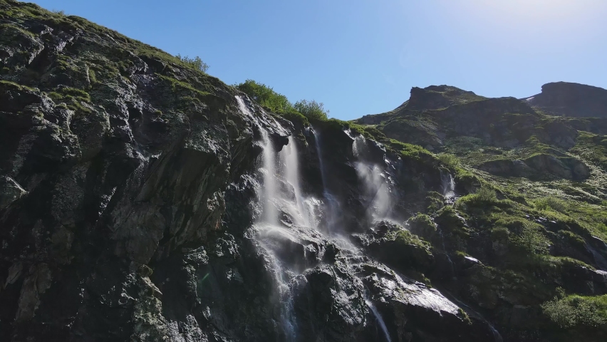 Water flows down the slope of a mountain covered with green vegetation, drops splash from a huge rock. Drops shimmer in the sun. Drone view. | Shutterstock HD Video #1091607391