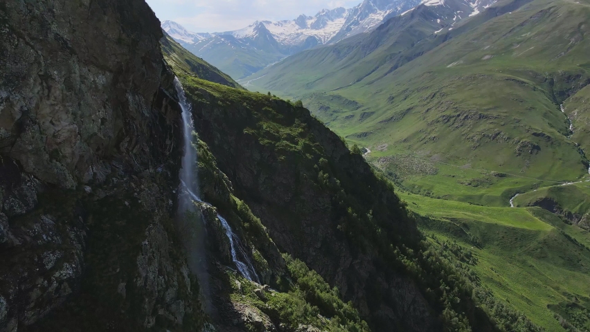 Water flows down the slope of a mountain covered with green vegetation, drops splash from a huge rock. Drops shimmer in the sun. Drone view. | Shutterstock HD Video #1091607399