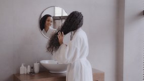 Young happy hispanic woman stands in bathroom in bathrobe looks to mirror combs thick long healthy curly hair using hairbrush feels satisfied morning daily routine advertise haircare treatment concept - Powered by Shutterstock - Get 15% off with code: PIKWIZARD15
