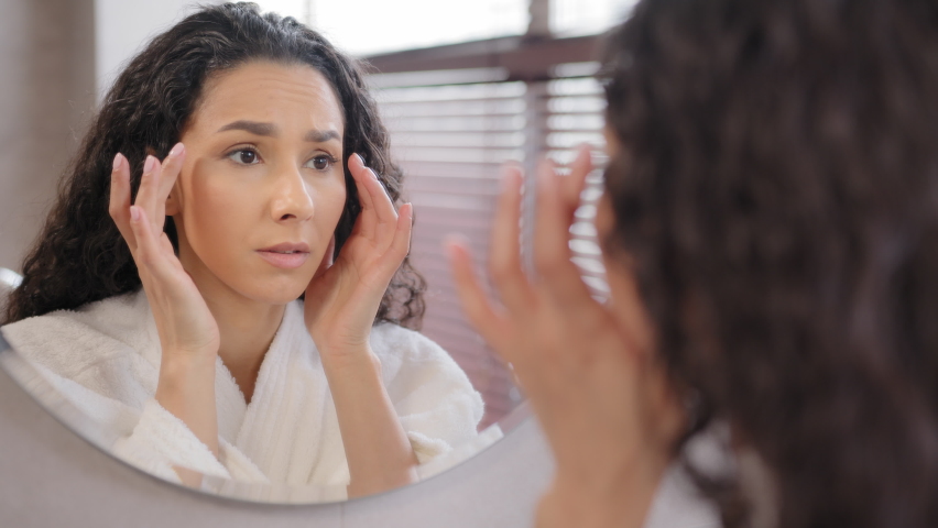 Upset worried young hispanic woman standing in bathroom looking in mirror worries about first wrinkles sad girl unhappy with condition of skin on face frustrated bad skincare product feeling stress Royalty-Free Stock Footage #1091609305