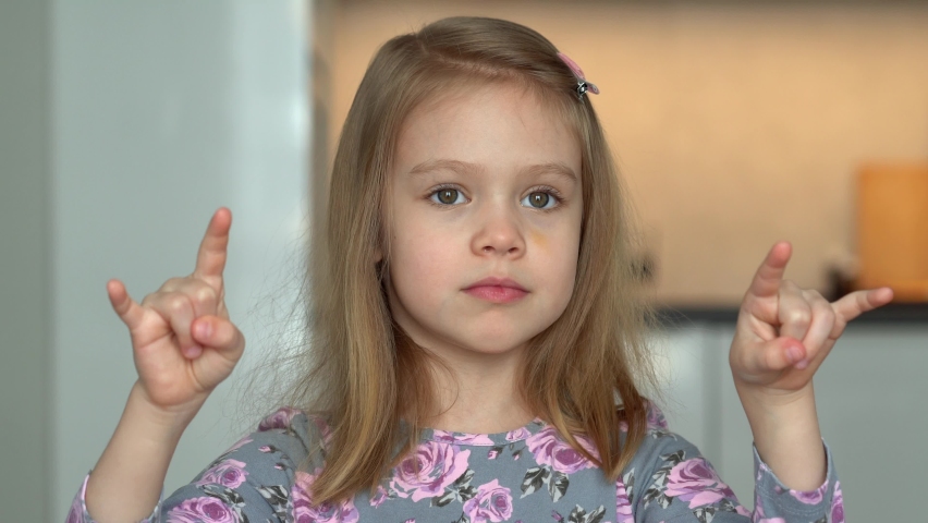 Portrait of little girl laughs and shows gesture with hands and fingers. Happy child looks at camera and plays with hands.