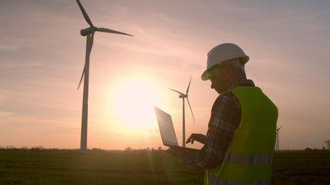 Man engineer works in the field of wind turbines. Electrical engineer working energy industry observing condition power equipment wind farm sunset. Checking data measurement results using laptop.