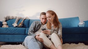 Couple Use Smartphone Device, while Sitting on a Couch in the Cozy Apartment. Boyfriend and Girlfriend Shopping on Internet, Watching Funny Videos, Use Social Media, Streaming Service. Medium Shot