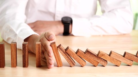 Risk management idea during a business crisis. A businessman's hand stops or blocks the effect of the risky dominoes falling onto a wooden table in slow motion.